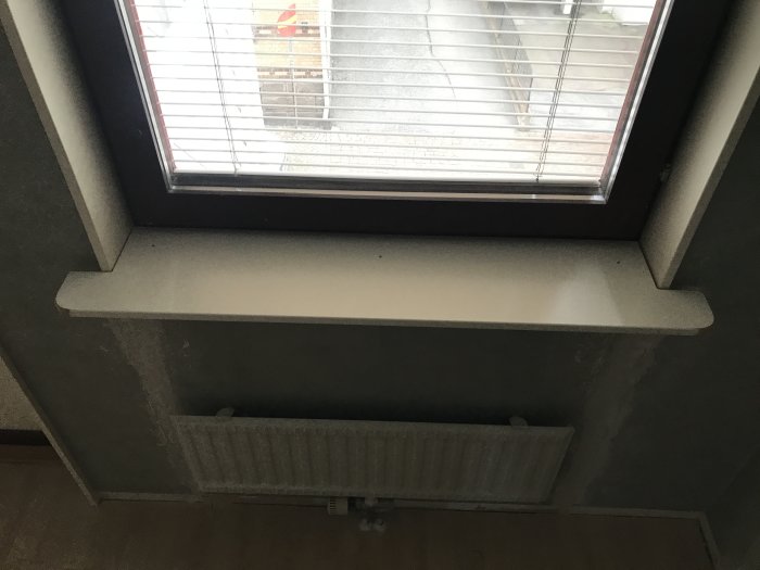 Infälld fönsterbräda ovanför en vit radiator under ett öppningsbart fönster med persienner.