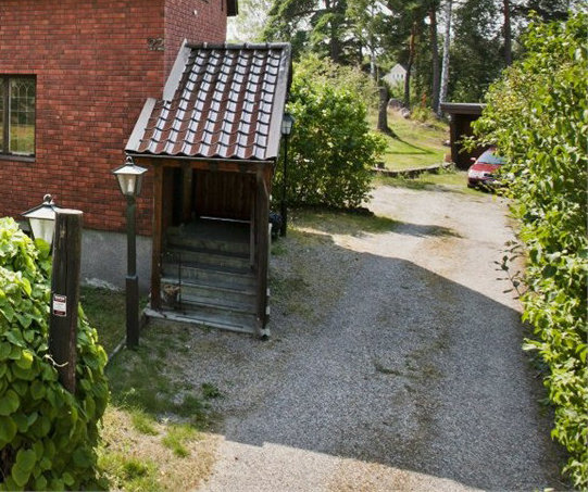 Gamla, slitna trätrappan vid husentré med tak, omgiven av grusgång och grönska.