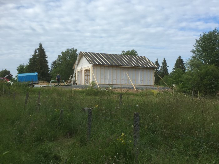 Byggnadsstomme i trä med pågående arbete och råspont på taket omgiven av grönt landskap.