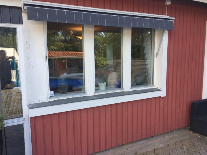 Nyligen installerat fönster på rödmålad fasad med vit listning och markis, reflektion av trädgården synlig.