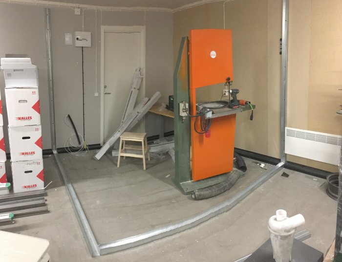 Inledande uppbyggnad av kontor med plåtreglar, verktyg och en stor orange maskin i ett snickeri under renovering.