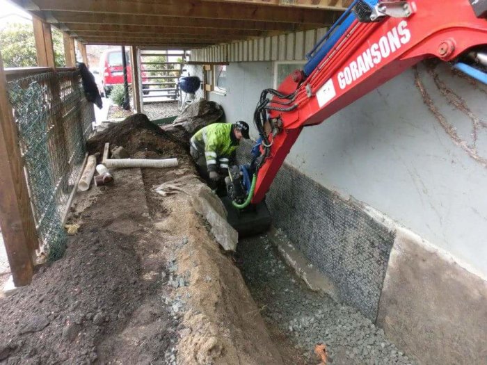 Arbetare tunnelgräver med en liten grävmaskin under en altan, omgiven av grushögar och material.