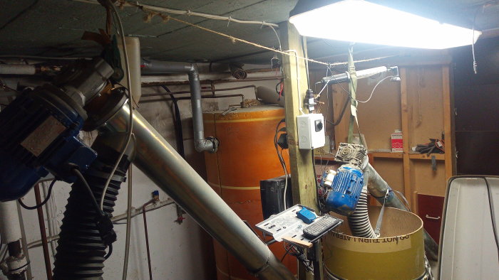 Rörledningar och maskinutrustning i ett trångt pannrum med en orange brännare och arbetslampor.