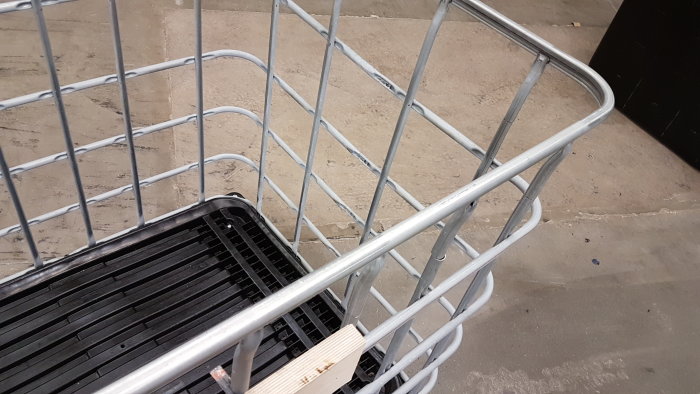 Metallbehållarram utan hål synlig ovanpå en svart plastpall på en betonggolv.