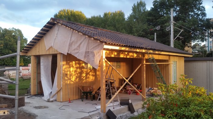 Hus under konstruktion med halvlagt tak av tegelpannor och byggmaterial synligt på marken vid skymning.