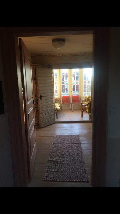 Utsikt från en öppen dörr mot en hall med vedpanelförsett tak och väggar, mönstrad matta på golv, och stolar mot fönster.