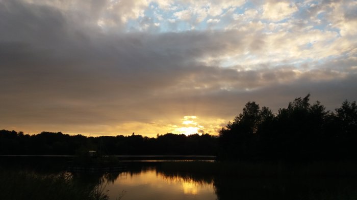 Solnedgång över en stilla sjö med spegelblank yta och träd vid horisonten, möjligt sista kvällsdopp.