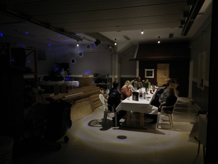 Oinredd verkstad/garage med människor runt bord och byggmaterial, med början av festbelysning.