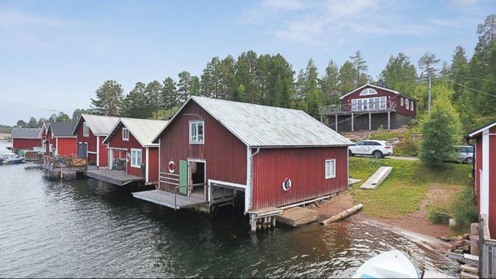 Röda sjöbodar på pålar vid vattnet med hus och bilar i bakgrunden.