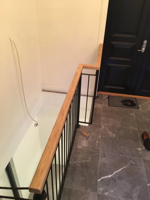 Obehandlad ekhandledare monterad på ett svart metallräcke vid en trappa, i en hall med marmorgolv och blå dörr.