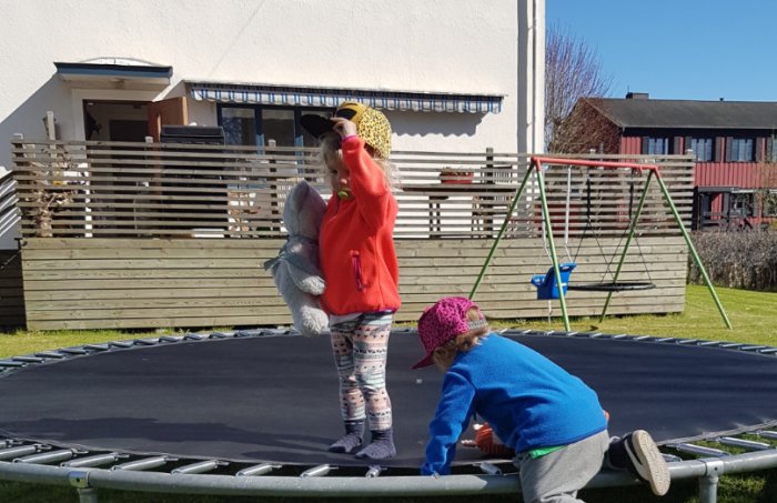 Barn leker på trampolin med en trädgårdsaltan och hög beväxtlighet i bakgrunden på en solig dag.