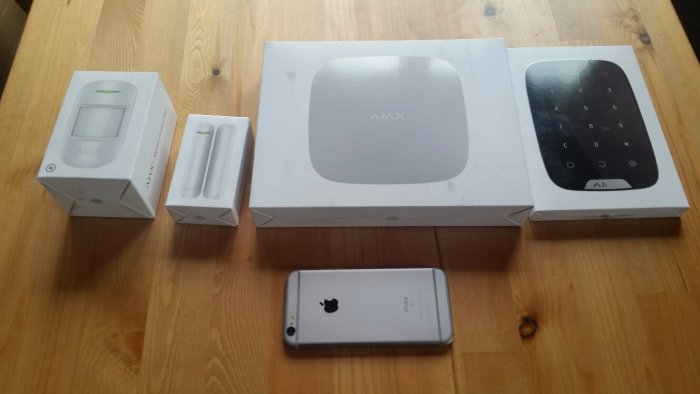 Ajax hemlarmssystem med sensorer och knappsats i förpackningar, bredvid en iPhone för storleksjämförelse.