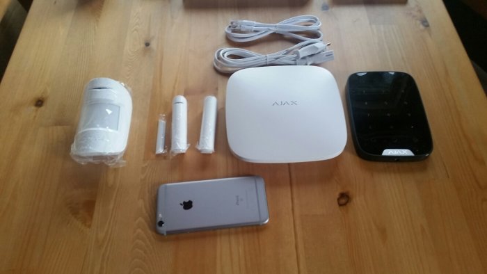 Ajax hemlarmssystemets komponenter uppackade på träbord, inklusive detektorer, kablar och en iPhone för storleksjämförelse.