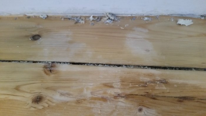 Trägolv med skrapade fläckar och färgrester nära väggkanten efter borttagning av färg från element.
