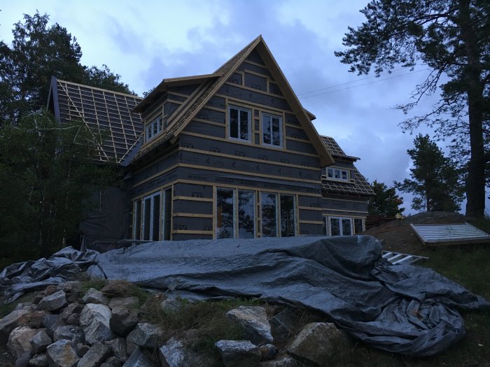 Nybyggt hus i träkonstruktion med många installerade fönster och dörrar, omgivet av sten och skog vid skymning.