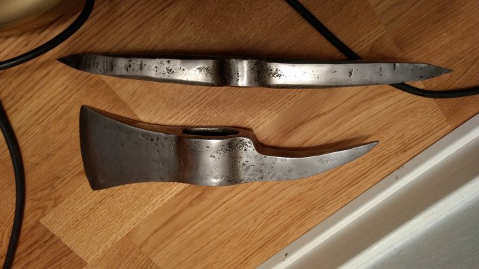 Två obehandlade metallväggprydnader i form av ett svärd och en yxa ligger på ett trägolv, i väntan på skaft och polering.