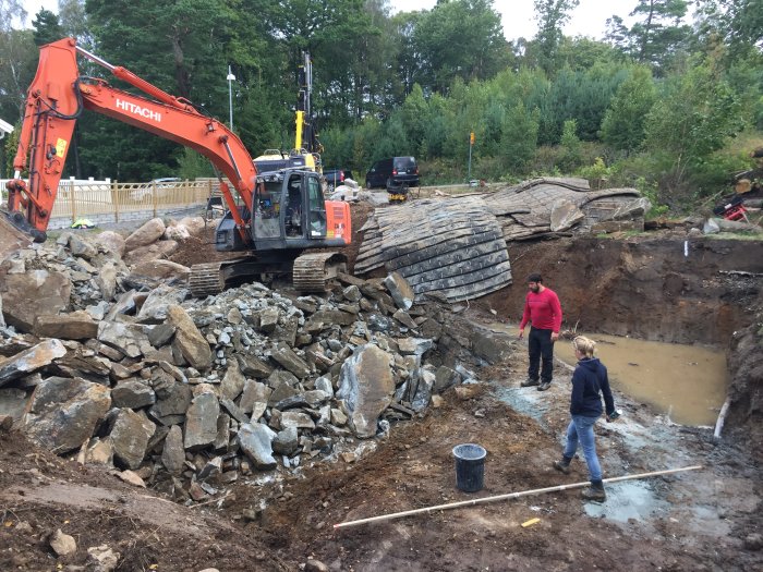 Efter sprängning: Grävmaskin, sprängsten och personer inspekterar en utgrävd byggplats.