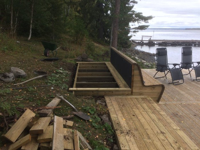 Uppbyggnad av utelkök bakom soffa på trädäck nära vatten med verktyg och byggmaterial.