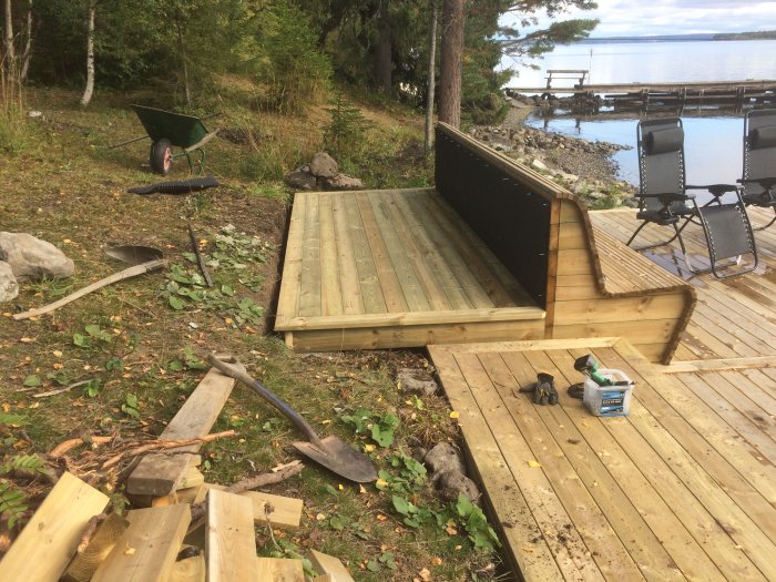 Pågående bygge av utomhuskök bakom en soffa på en trädäcksväranda nära en sjö, med verktyg och byggmaterial.