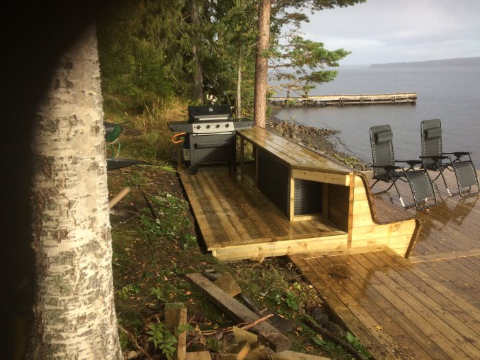 Utelkök av trä bakom soffa på träaltan med utsikt över sjö och brygga.