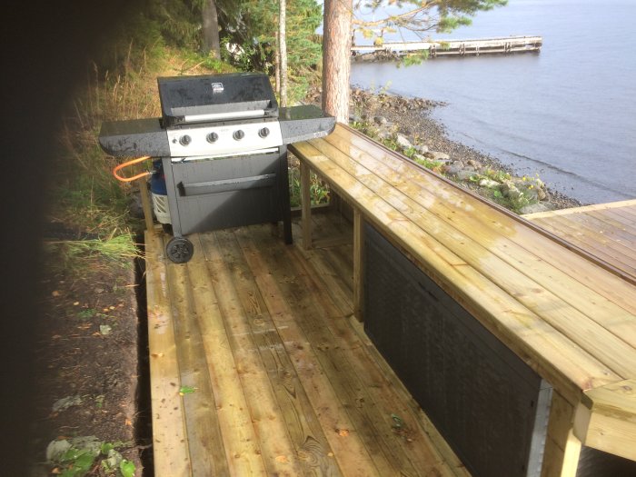 Ett utelkök med gasolgrill på en trädäck med sjöutsikt; plats för förvaring under bänken till höger.