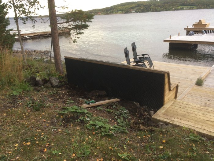 Pågående bygge av ett utelkök bakom en soffa på en träaltan vid en sjö med brygga i bakgrunden.