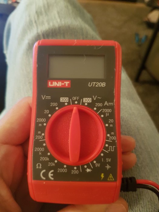 En hand håller en UNI-T UT20B multimeter med röd vred mot en bakgrund av jeansklädda ben.