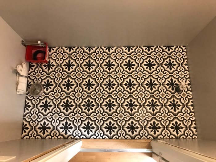Vägg med mönstrade kakelplattor och synligt rörarbete samt hål för toalett och avlopp.