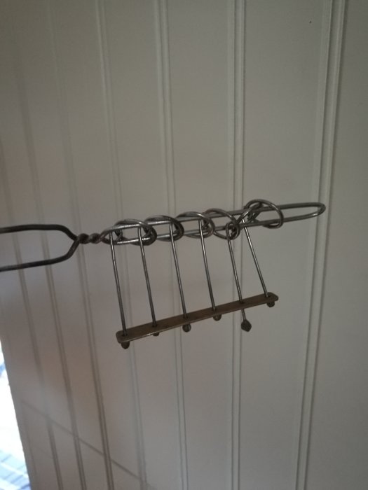 Ett okänt hängande metallföremål med krokar och trädetalj mot en vit panelvägg.