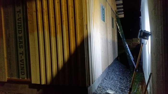 Målning av träpanel på en husvägg i dunkel belysning från en arbetslampa, med duggregn och mörker omkring.