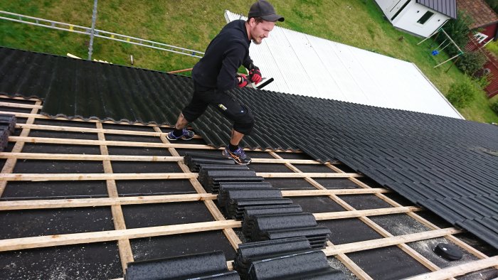 Person som lägger svarta takpannor på träkonstruktion på ett hustak.