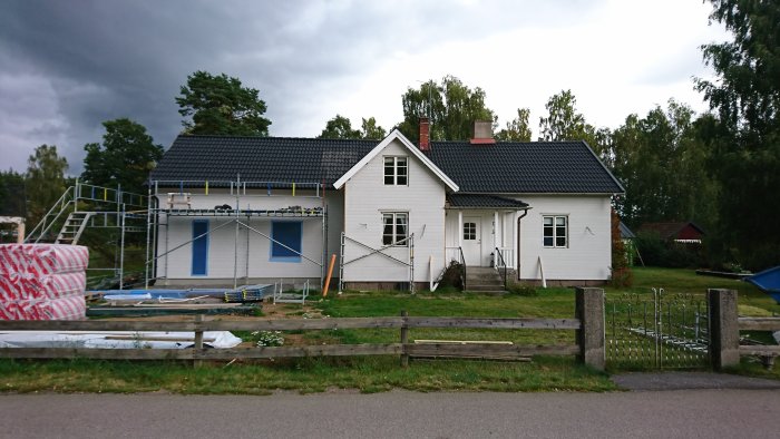 Ett vitmålat hus under renovering med nya svarta takpannor och byggställning på sidan.