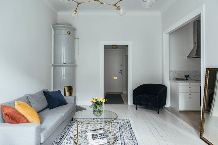 Renoverat vardagsrum med grå soffa, glasbord, blå fotölj och vit kakelugn, angränsande till ett modernt kök.