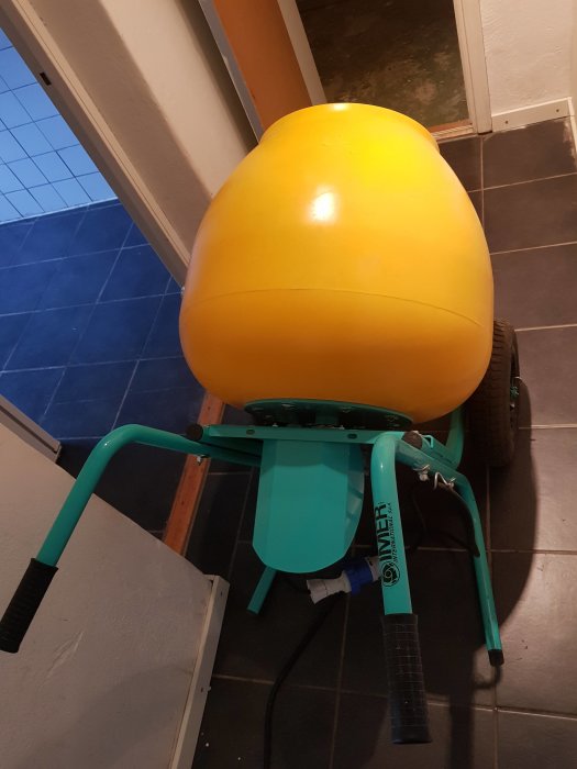 Gul och grön Rollbeta betongblandare på ett blått kakelgolv med en öppen dörr i bakgrunden.