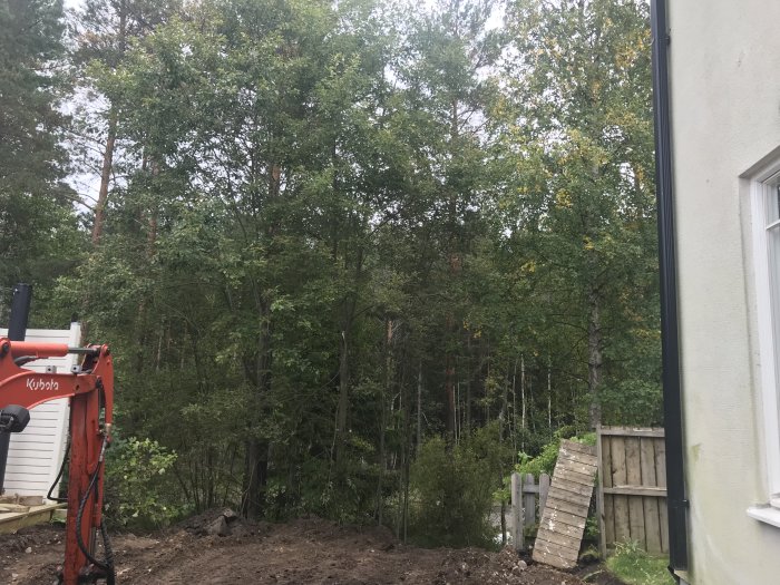 Grävmaskin vid grävt område nära hus med utsikt över skog och staket.