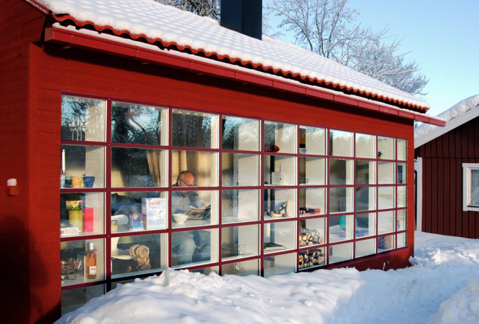 Stor fönstervägg med många spröjs på ett rött hus, snötäckt tak, i vintersol, reflekterar exempel på modern tillbyggnad.