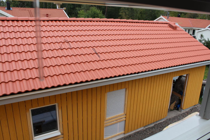 Nästan färdigt tak med röda tegelpannor och en monterad fönsterruta på en gul trähusfasad.
