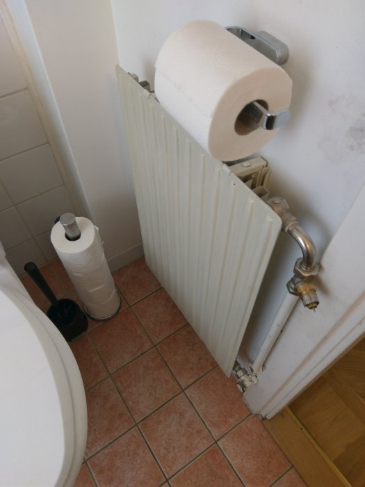 En vit enkelpanel radiator i ett badrum med röranslutningar och toalettpapper ovanpå.