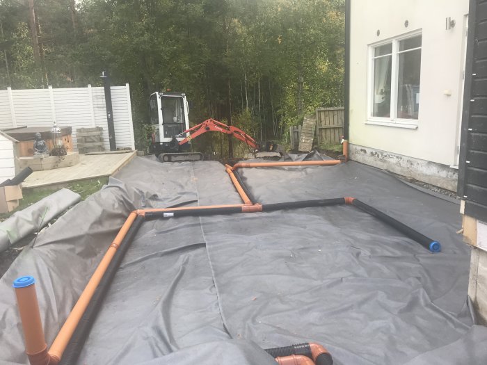 Uppgrävd trädgård med dräneringsrör och markduk vid en villa med en grävmaskin i bakgrunden.