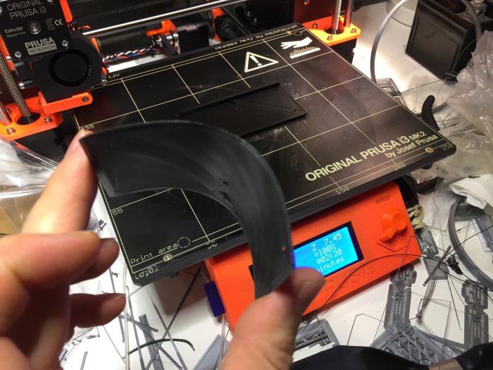 Hand som böjer ett svart 3D-utskrivet objekt framför en Prusa 3D-skrivare.