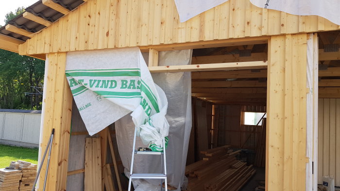 En byggarbetsplats med träkonstruktion, reglar och vindduk ovanför en port, med byggmaterial i förgrunden.
