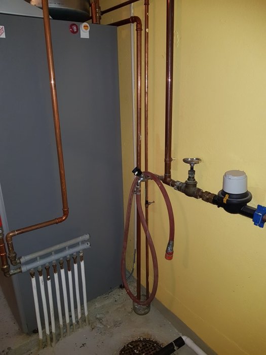 VVS-installation med spillrör från säkerhetsventil, rör i väggen, radiator och manometer i ett pannrum.