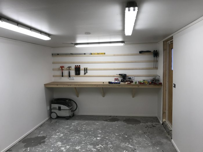 En verkstad med en arbetsbänk och "French cleat" verktygstavla med några verktyg på, samt dammsugare i hörnet.