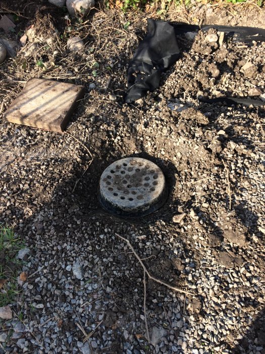 Uppgrävd uppfart med synligt borrhål för bergvärme och skevt plastlock omgivet av grus och jord.