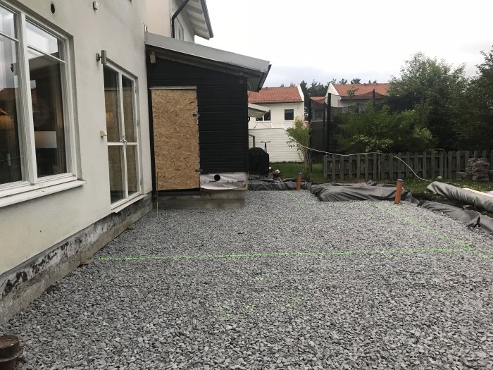 Grusad yta för blivande altan markerad med gröna linjer bredvid ett hus med plywoodförsluten dörr.