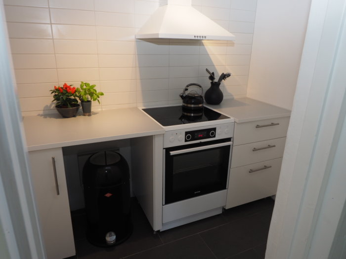 Renoverat kök med vit spis och köksfläkt, vita skåp och kakel, samt två krukväxter på bänkskivan.