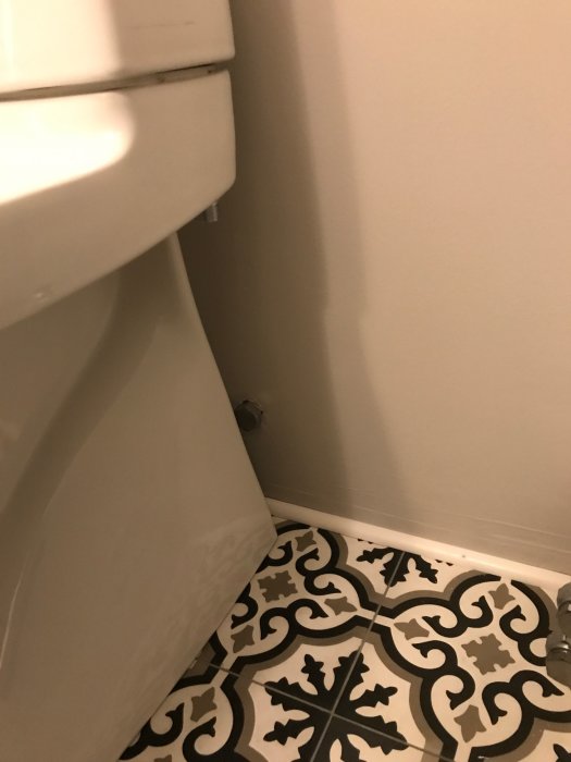 Toalettstol nära väggen med vattenanslutning bakom, ojämnt placerad på ett mönstrat golv.
