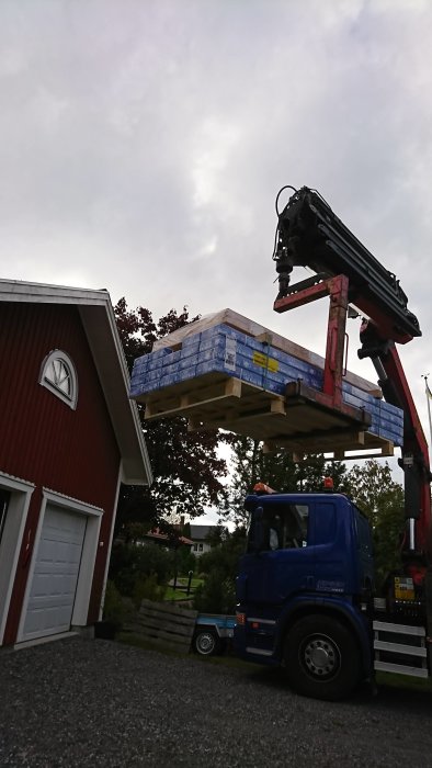 Lastbil med kran lyfter byggmaterial till ett hus under en molnig himmel.