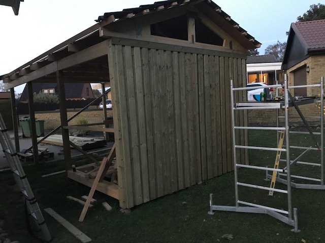 Uppförandet av en liten träbyggnad med nyspikad panel, delvis täckt tak och arbetsstege i förgrunden.