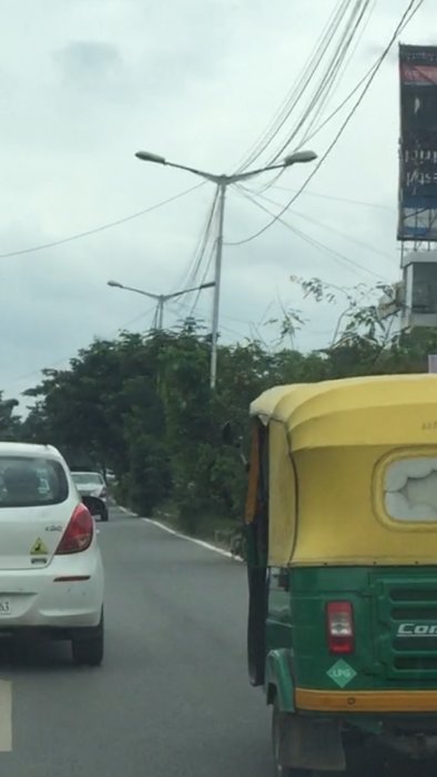 En gata i Indien med trassliga fiberkablar hängandes över belysningsstolpar, framför träd och vid sidan av en auto-rickshaw.